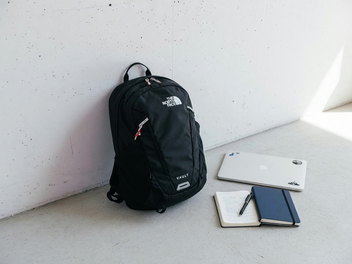 Minimalist black North Face Vault backpack leaning against a white concrete wall beside a laptop and notebook, bright
